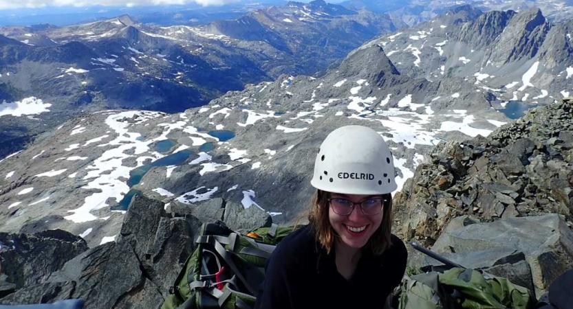 A person wearing a helmet smiles while standing above snow capped mountains. 